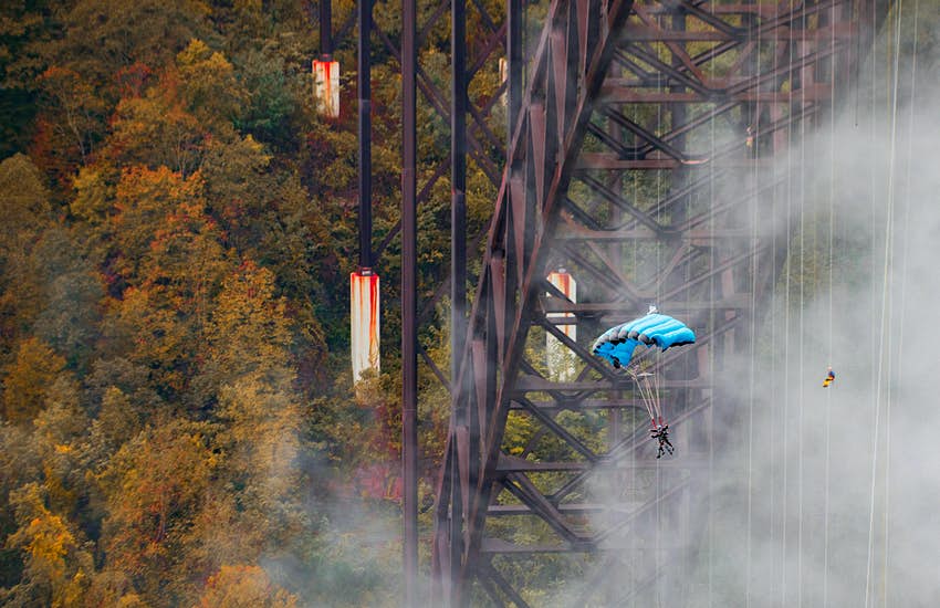 Dos personas sujetas a un paracaídas se deslizan bajo el puente New Gorge durante el día.  El bosque y las vigas de soporte del puente son visibles.